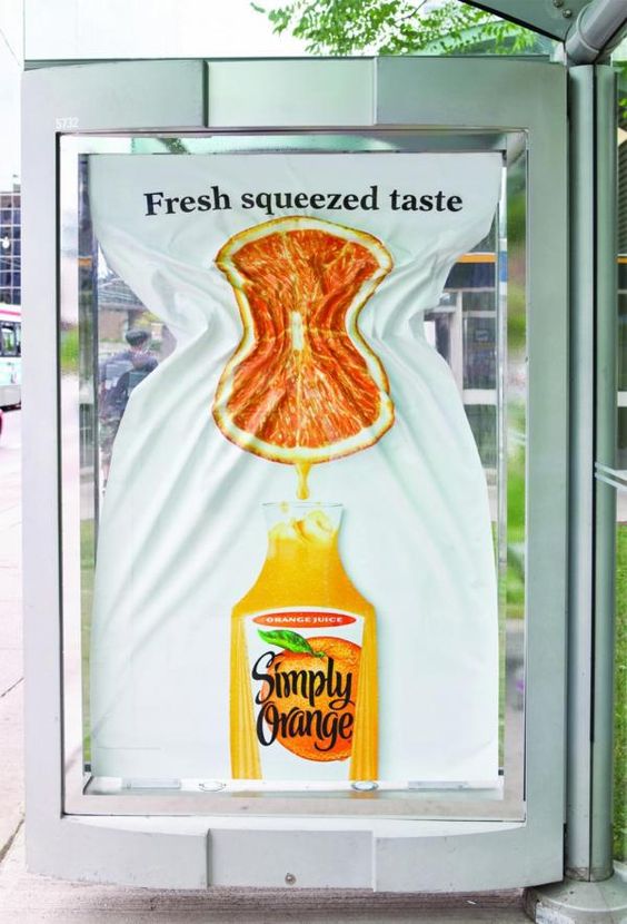 Campanha da empresa Simply Orange. Na foto está uma parada de onibus onde a parte de cima do banner está espremendo uma laranja, abaxio está a garrafa com o suco da empresa