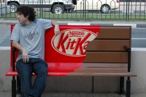 Camoanha de Marketing de Guerrilha da Nestle. Na foto temos homem sentado em um banco todo marrom em volta de uma embalagem de Kitkat.
